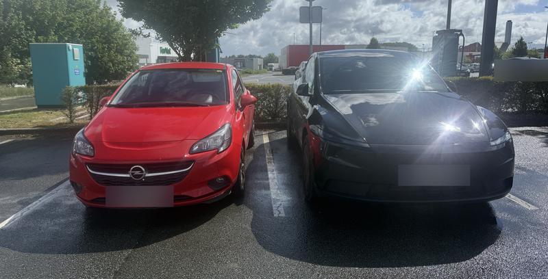 Tesla Model Y noire garée à côté d’une citadine thermique rouge sur un parking